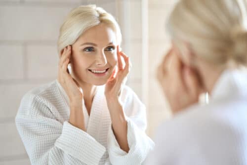 mid age adult 50 years old blonde woman standing in bathroom after shower touching face 1 1