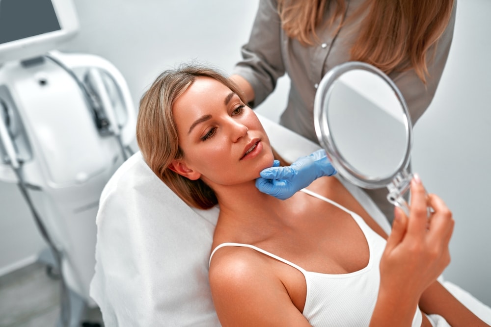 Cosmetologist consulting a female client in a beauty clinic as she looks in the mirror, preparing for a skincare procedure such as SmartXide DOT laser resurfacing for skin rejuvenation.