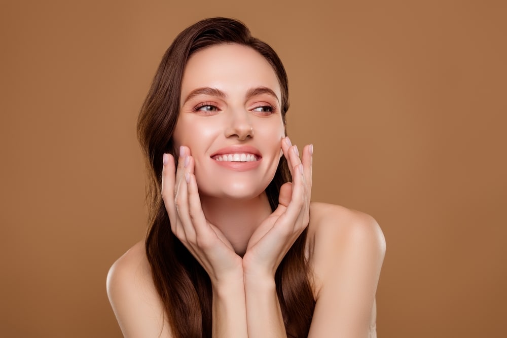 Smiling woman touching cheeks while applying facial peeling cream, concept of skin rejuvenation and neuromodulators.